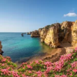 A stunning coastal view of the Algarve region in Portugal, showcasing towering golden cliffs and rock formations rising from the turquoise waters of the Atlantic Ocean. Vibrant pink flowers frame the foreground, while a secluded sandy beach nestles between the cliffs under a clear blue sky.