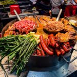 A vibrant street food scene in Mexico City featuring a large round griddle filled with various meats, including chorizo sausages, beef, and pork, along with fresh green onions. The sizzling food is being prepared by cooks wearing white shirts and red aprons in the background, while tortillas are being heated on a separate grill. The rich colors and textures highlight the authentic flavors of traditional Mexican cuisine.
