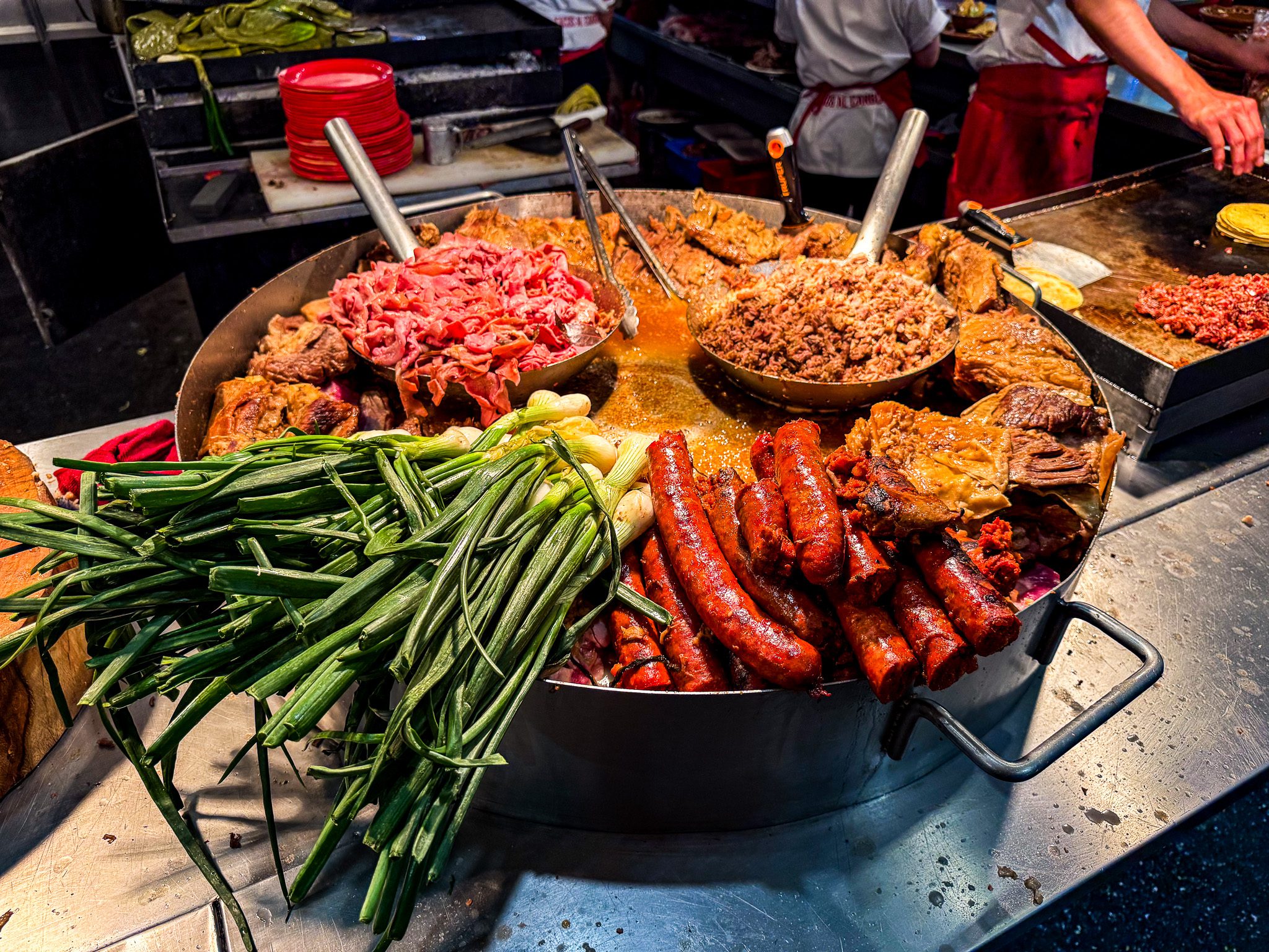 A vibrant street food scene in Mexico City featuring a large round griddle filled with various meats, including chorizo sausages, beef, and pork, along with fresh green onions. The sizzling food is being prepared by cooks wearing white shirts and red aprons in the background, while tortillas are being heated on a separate grill. The rich colors and textures highlight the authentic flavors of traditional Mexican cuisine.