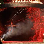 A person ziplining at night, arms outstretched, passing through a stone arch engulfed in flames, with trees and red lighting creating a dramatic backdrop.