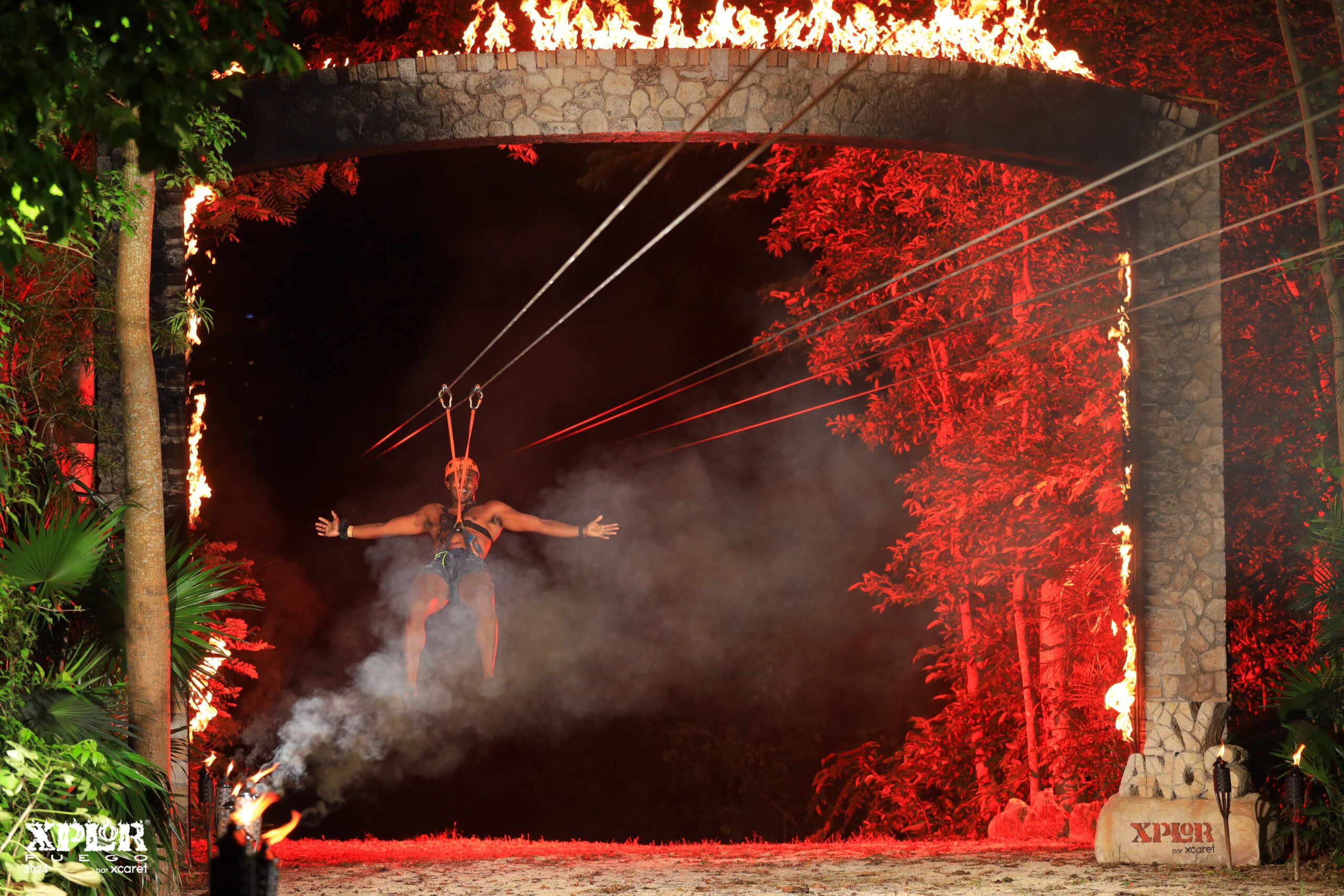 A person ziplining at night, arms outstretched, passing through a stone arch engulfed in flames, with trees and red lighting creating a dramatic backdrop.
