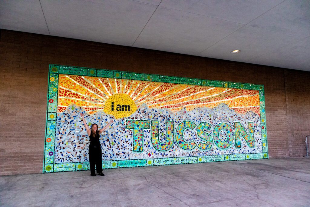Woman standing with arms raised in front of a colorful mosaic mural that reads 'I am Tucson'