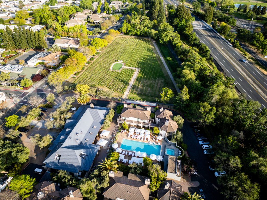 Bird’s-eye view of The Estate Yountville, showing the hotel grounds, pool area, and adjacent vineyard with nearby Yountville streets and highway in the background.