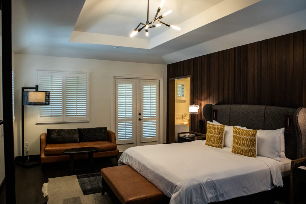 Luxury guest room at The Estate Yountville with king bed, leather seating area, modern chandelier, and French doors leading to a private patio
