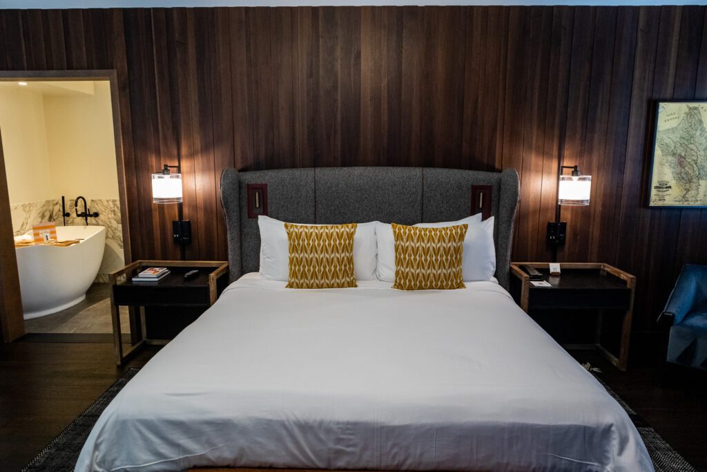 Elegant hotel room at The Estate Yountville featuring a king-size bed with yellow accent pillows, wood-paneled walls, and a view of the freestanding bathtub in the marble bathroom