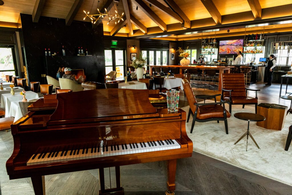 Warm and stylish lobby bar at The Estate Yountville featuring a K. Kawai grand piano, leather chairs, and a modern chandelier.