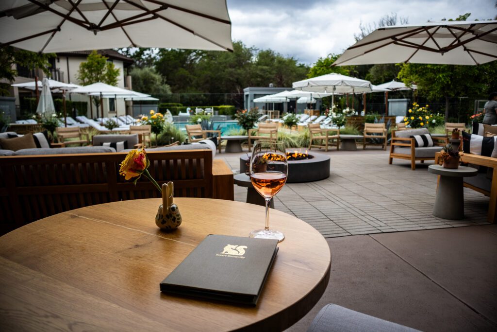 Outdoor fire pit lounge at The Estate Yountville with a glass of rosé, surrounded by cushioned chairs and umbrellas near the pool.