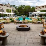 Central pool area at The Estate Yountville with lounge chairs, white umbrellas, lush landscaping, and a round modern fire pit.