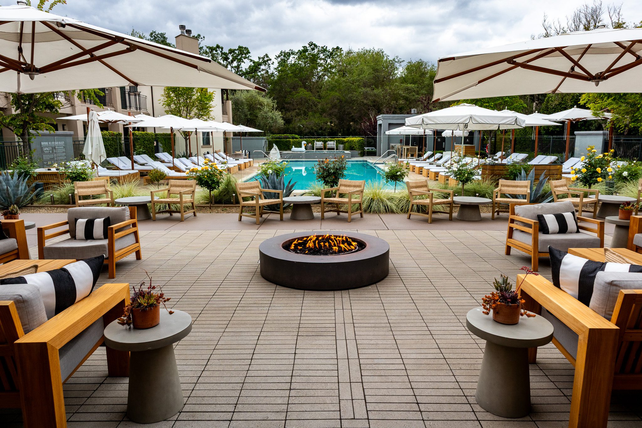 Central pool area at The Estate Yountville with lounge chairs, white umbrellas, lush landscaping, and a round modern fire pit.