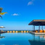 A serene infinity pool overlooking the ocean under a clear blue sky, with a single palm tree on the left, white umbrellas shading lounge chairs, and a modern open-air pavilion with cushioned seating on the right.