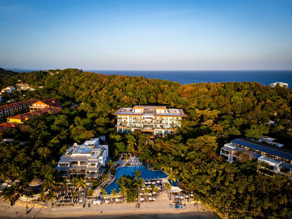 Aerial view of Kimpton Grand Roatan surrounded by lush jungle, with the resort’s pool and beachfront area in the foreground and the Caribbean Sea in the distance.