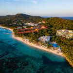 Aerial photo of Kimpton Grand Roatan resort nestled between turquoise water and dense greenery, showing beachfront access, pool, and surrounding buildings.