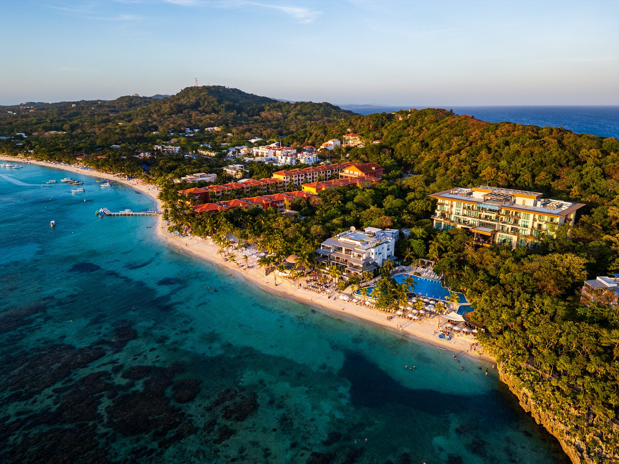 Aerial photo of Kimpton Grand Roatan resort nestled between turquoise water and dense greenery, showing beachfront access, pool, and surrounding buildings.