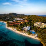 Aerial view of Kimpton Grand Roatan surrounded by lush jungle, with the resort’s pool and beachfront area in the foreground and the Caribbean Sea in the distance.