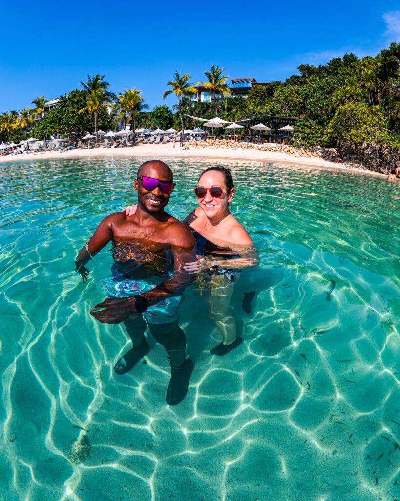 Smiling couple standing in crystal-clear turquoise water in front of Kimpton Grand Roatan, with palm trees, beach loungers, and umbrellas lining the sandy shore behind them.