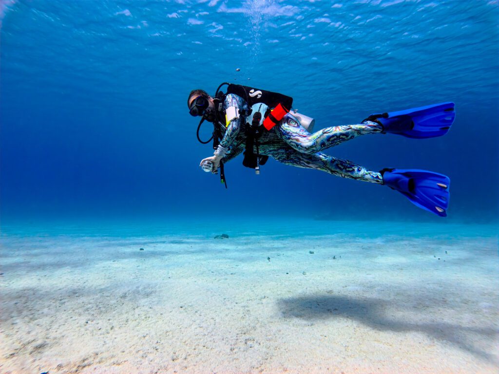 Scuba diver floating above the sandy ocean floor in crystal-clear blue water, fully geared with blue fins and patterned dive suit in Roatan.