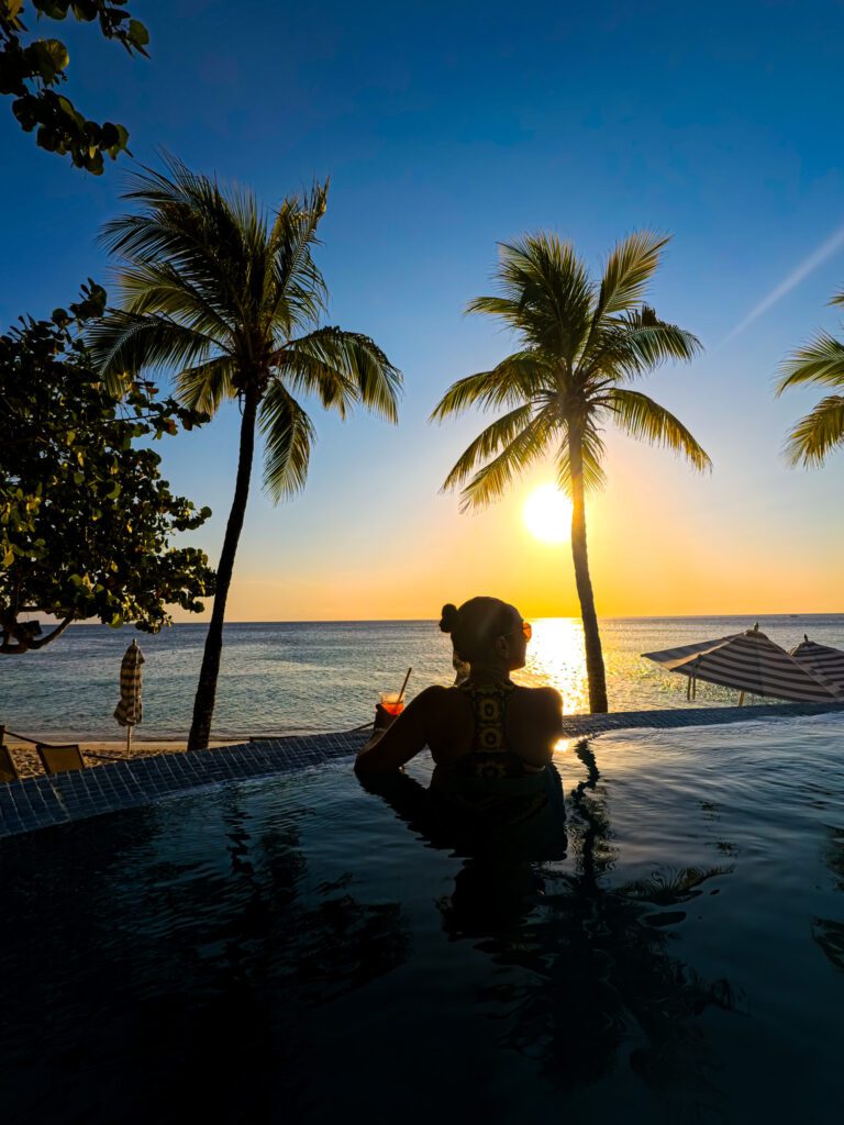 Woman enjoying a cocktail in an infinity pool at sunset, gazing at the ocean with silhouetted palm trees and golden sunlight reflecting off the water.