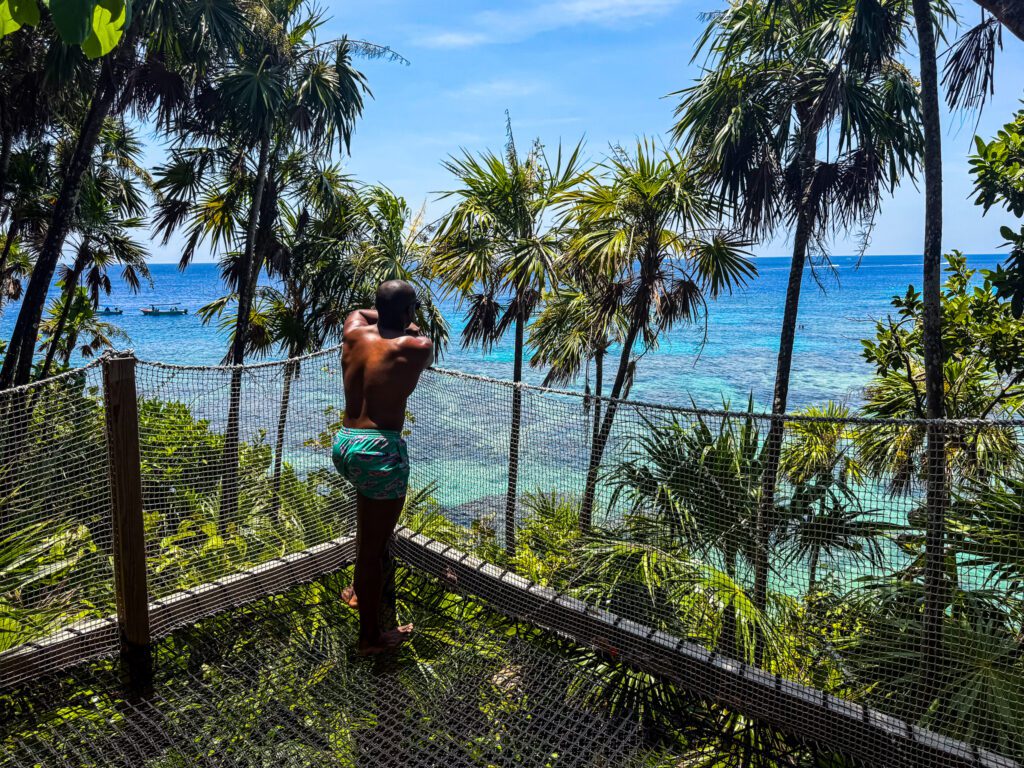 Man standing shirtless on a jungle viewing platform overlooking turquoise Caribbean waters, surrounded by palm trees on a sunny day.