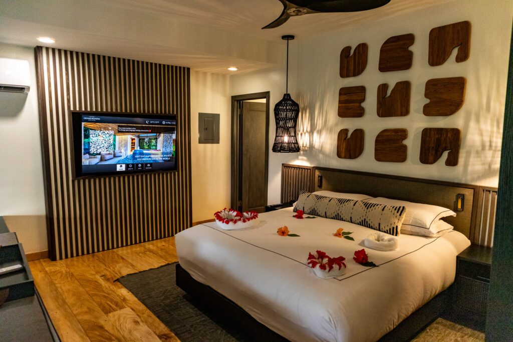Beautifully prepared king bed at Kimpton Grand Roatan decorated with hibiscus flowers and towel swans, with modern decor and a welcome message on the TV screen.