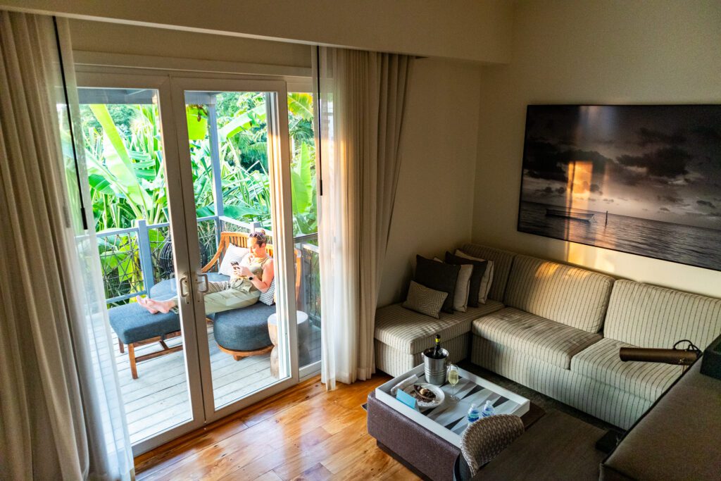 Interior view of a Kimpton Grand Roatan bungalow suite, looking out onto a private jungle-facing balcony where a woman relaxes with a drink.