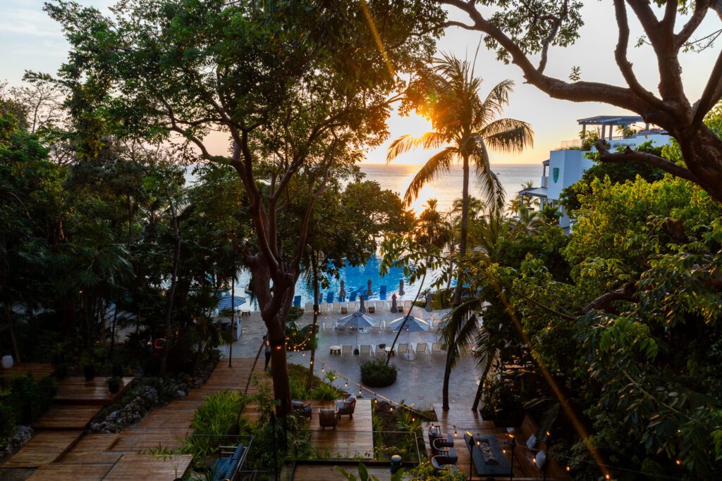 View through tropical trees at sunset from Kimpton Grand Roatan, overlooking the resort’s pool area and the Caribbean Sea beyond, with string lights and wooden walkways adding ambiance.
