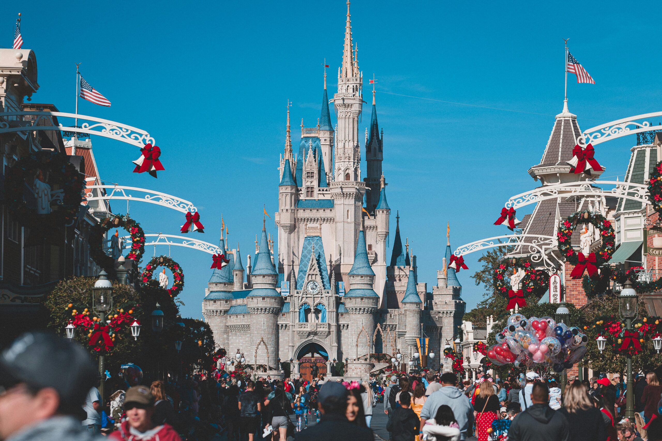 Crowds of visitors walk down Main Street at Disney World decorated with holiday wreaths and red bows, leading toward Cinderella Castle under a clear blue sky.