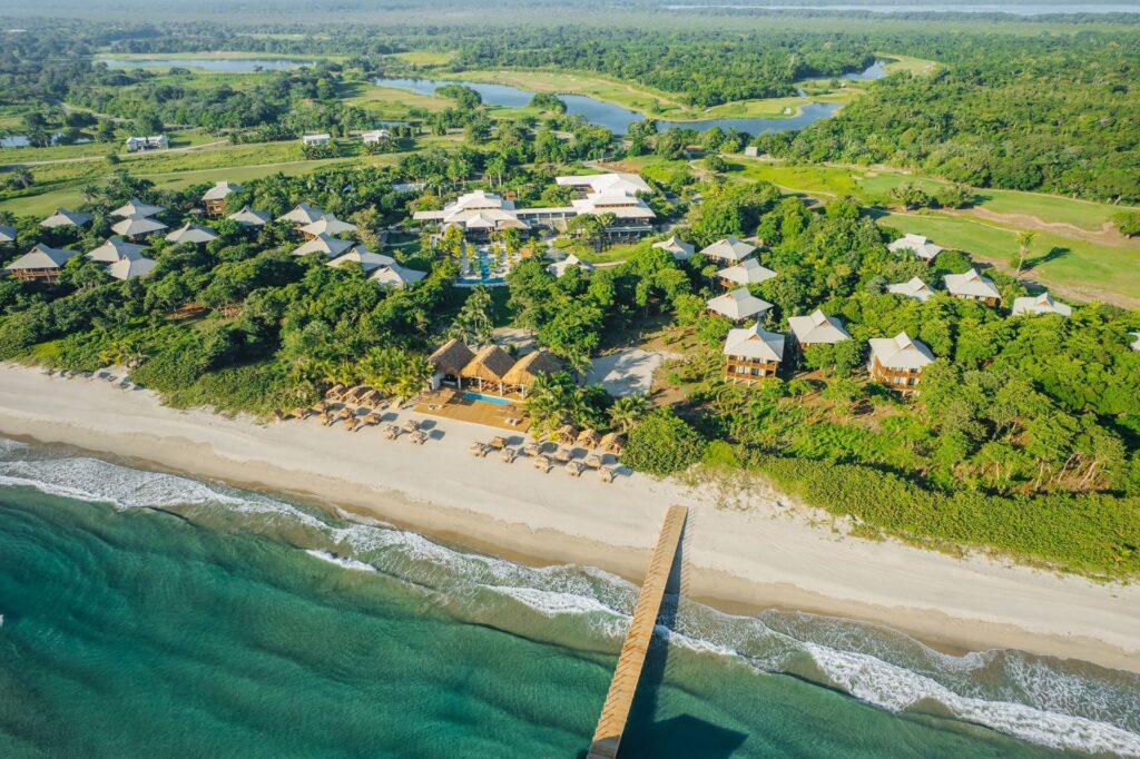 Aerial view of Indura Beach & Golf Resort in Tela, Honduras, showing beachfront bungalows, a pier stretching into the Caribbean Sea, thatched palapas on the sand, and lush greenery with golf course in the background