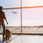 Man looking out airport window with his carry ons as a plane flies overhead in the distance