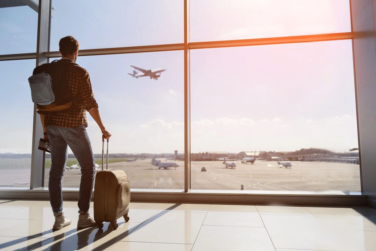 Man looking out airport window with his carry ons as a plane flies overhead in the distance