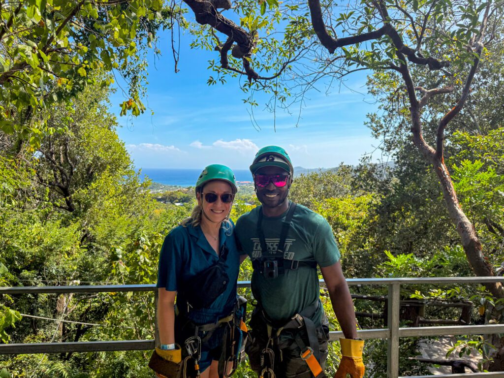A couple wearing helmets and zipline gear smile together with lush jungle greenery and ocean views in the background at Mayan Eden in Roatán.