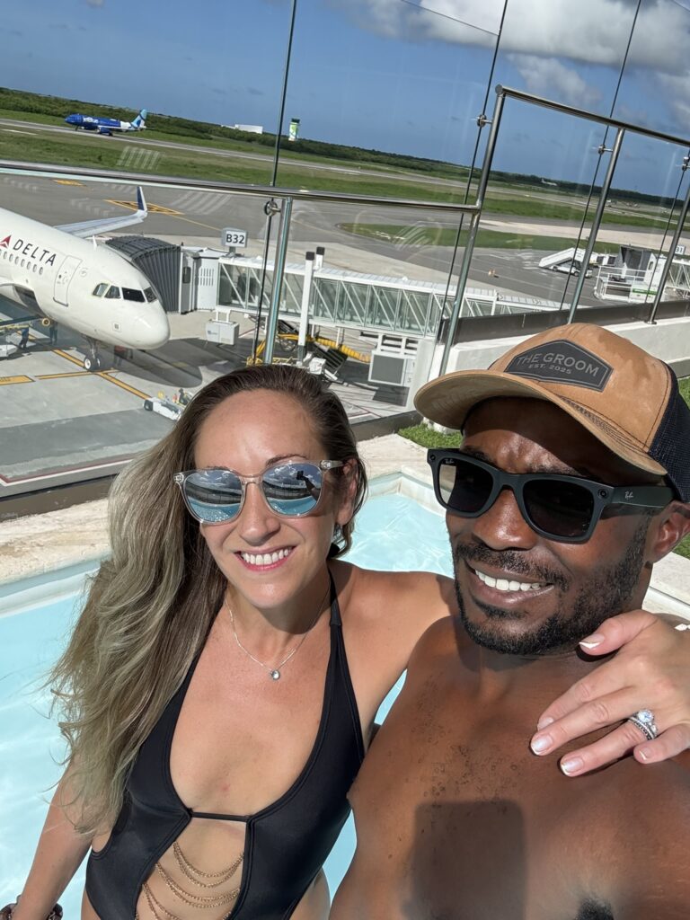 Couple enjoying the rooftop pool at the Punta Cana VIP Lounge with clear glass railings and airplanes visible on the tarmac below on a sunny day.