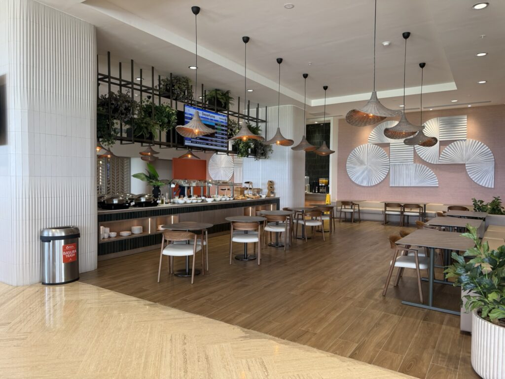 Buffet dining section at the Punta Cana Airport VIP Lounge with hanging pendant lights, modern wooden tables, and breakfast or lunch spread displayed along the counter.