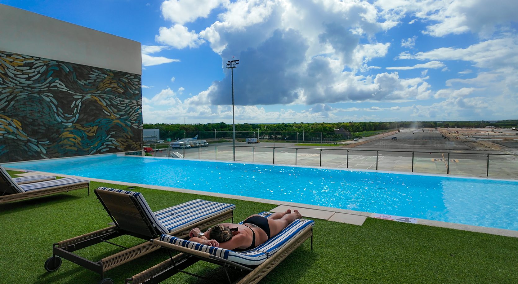 Infinity pool at the Punta Cana Airport VIP Lounge with striped loungers, modern mural wall art, and expansive runway views under bright tropical skies.