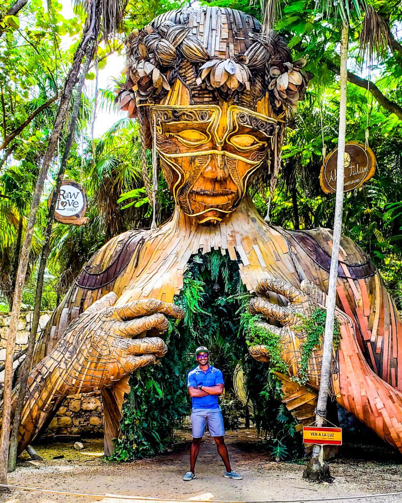 Man standing in front of the famous ‘Ven a la Luz’ wooden art installation in Tulum, a towering sculpture of a human figure opening its chest to reveal lush greenery.