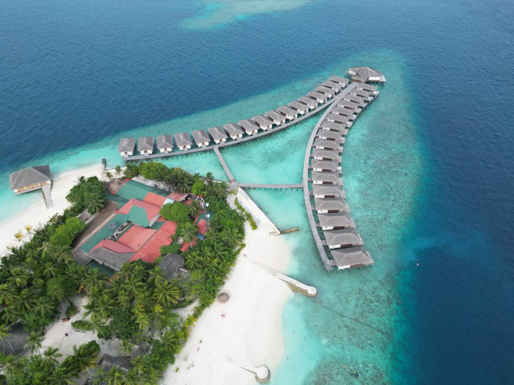 Aerial drone view of a Maldives island resort with overwater villas curving above turquoise lagoons, white-sand beaches, and lush palm trees surrounded by deep blue ocean.