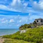 Ancient Mayan ruins sitting on a rocky cliff overlooking the turquoise Caribbean Sea in Tulum, Mexico, surrounded by lush green vegetation under a bright blue sky with scattered clouds.