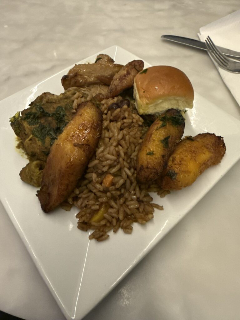 Plate of Jamaican dishes from the Regency Club’s Caribbean Night, including jerk chicken, rice and peas, plantains, and a dinner roll.