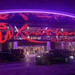 Wide view of the Resorts World New York City entrance with cars parked under bright purple lighting.