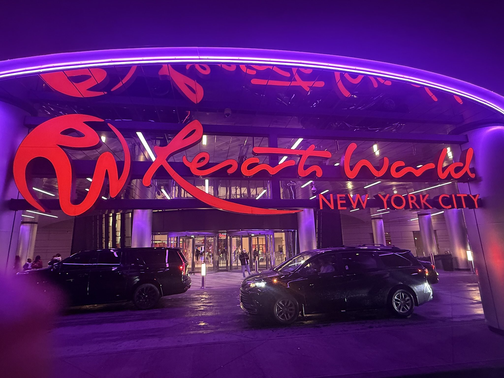 Wide view of the Resorts World New York City entrance with cars parked under bright purple lighting.