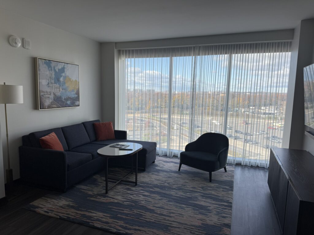 Executive Suite living room at Hyatt Regency JFK with large windows, natural light, sofa, armchair, and city views.