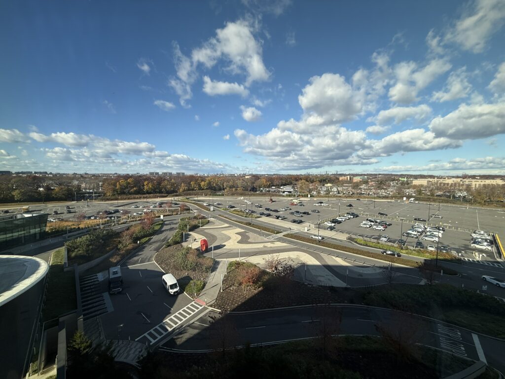 View from Hyatt Regency JFK overlooking parking areas, landscaped paths, and Queens skyline under a partly cloudy sky.