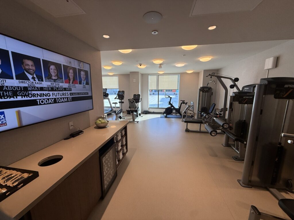 Hyatt Regency JFK fitness center with cardio machines, weight equipment, and a large TV wall.