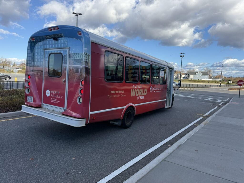 Red Resorts World New York City shuttle bus branded with Hyatt Regency JFK, parked outdoors under a partly cloudy sky.