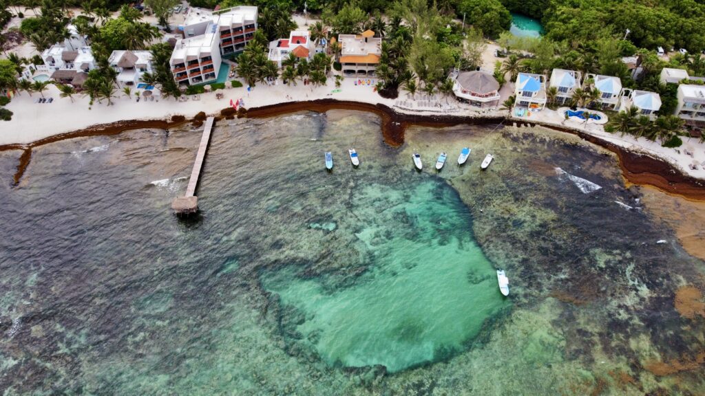 Aerial view of Tulum’s coastline with turquoise water, boats near the reef, beachfront hotels, and a line of sargassum along the shore.