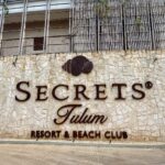 Exterior stone wall with the “Secrets Tulum Resort & Beach Club” sign and building façade above.