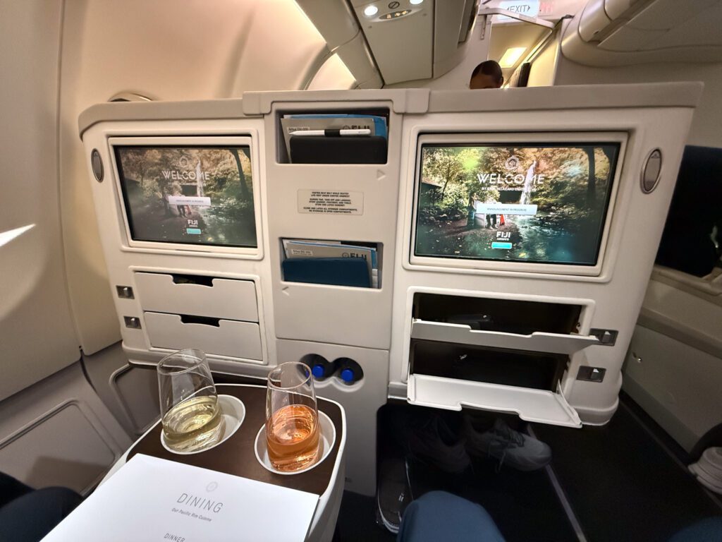 Front view of Fiji Airways A330 business class seat showing dual entertainment screens, limited storage compartments, and welcome screen display.
