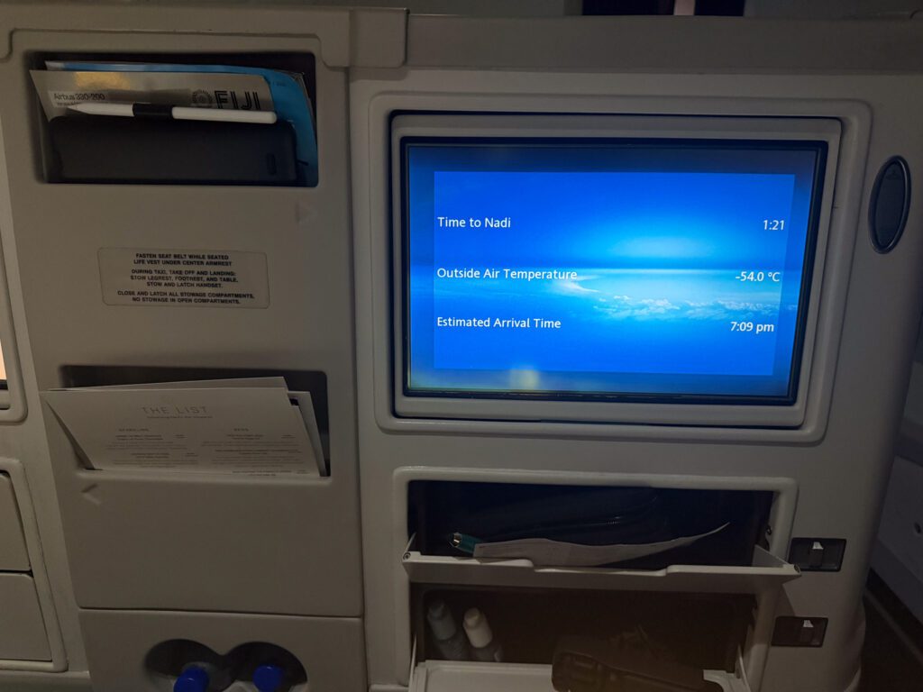 Fiji Airways A330 business class entertainment screen showing time to Nadi with visible seat storage compartments and limited personal space.
