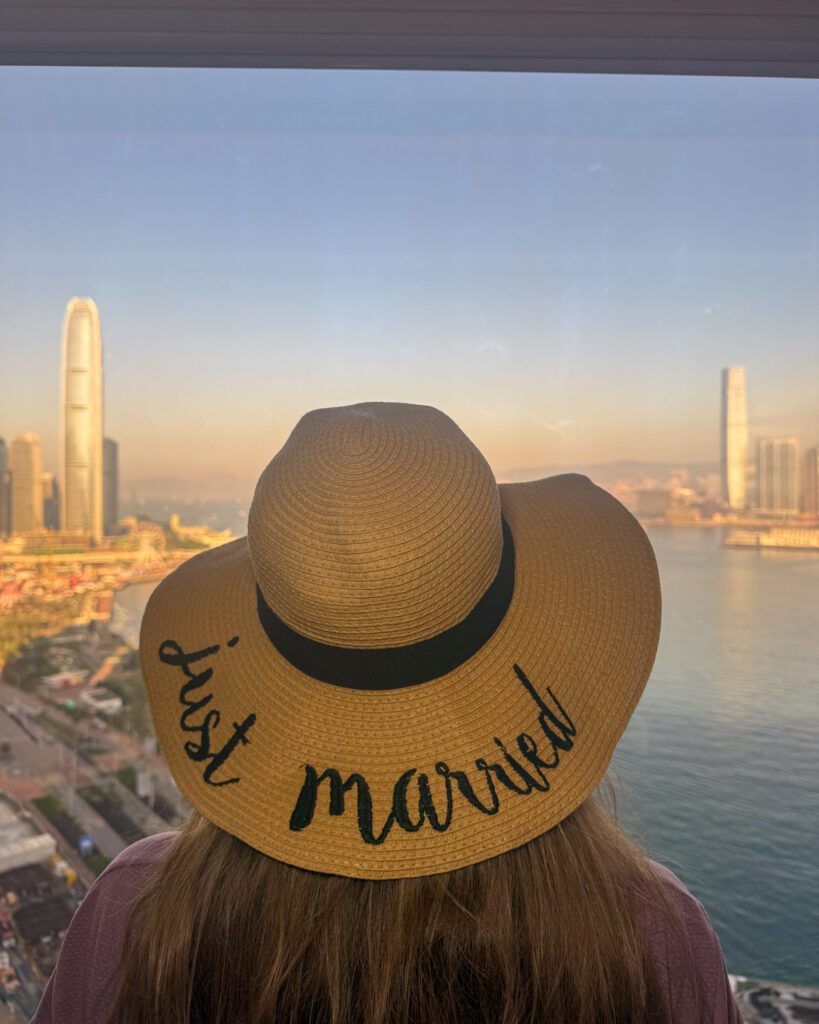 Traveler wearing a “Just Married” hat looking out at Victoria Harbour and the Hong Kong skyline at sunset