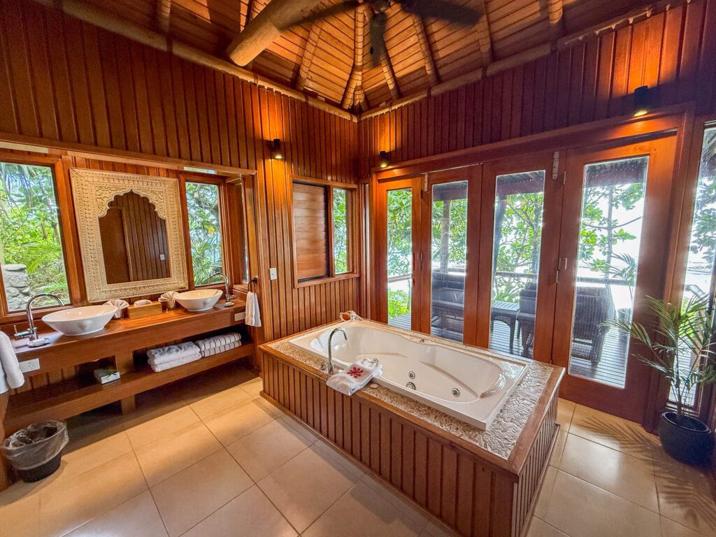 Luxury villa bathroom in Fiji with dual vessel sinks, soaking tub, and glass doors opening to tropical greenery.
