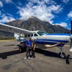 Couple standing in front of a small scenic plane in Milford Sound, New Zealand, with mountains and blue sky during a fly-cruise experience